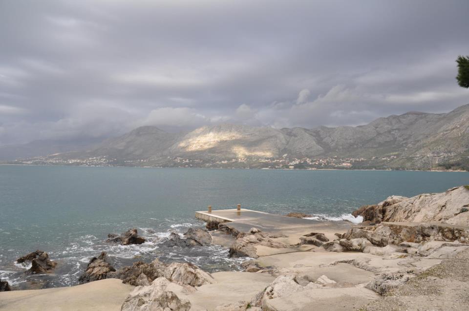 A scenic view of a calm sea with rocky shores under a cloudy sky, showcasing a distant coastline with mountains.