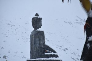 Stone Buddha statue in gentle snowfall, with snow-covered hills in the background.