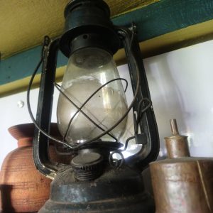 Close-up of an antique oil lantern with a glass chimney, surrounded by wooden and metallic objects on a shelf.