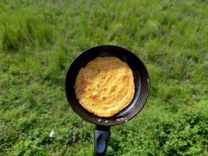 A golden-brown flatbread (roti/chapati) freshly cooked in a small black pan, held up against a lush green meadow. 