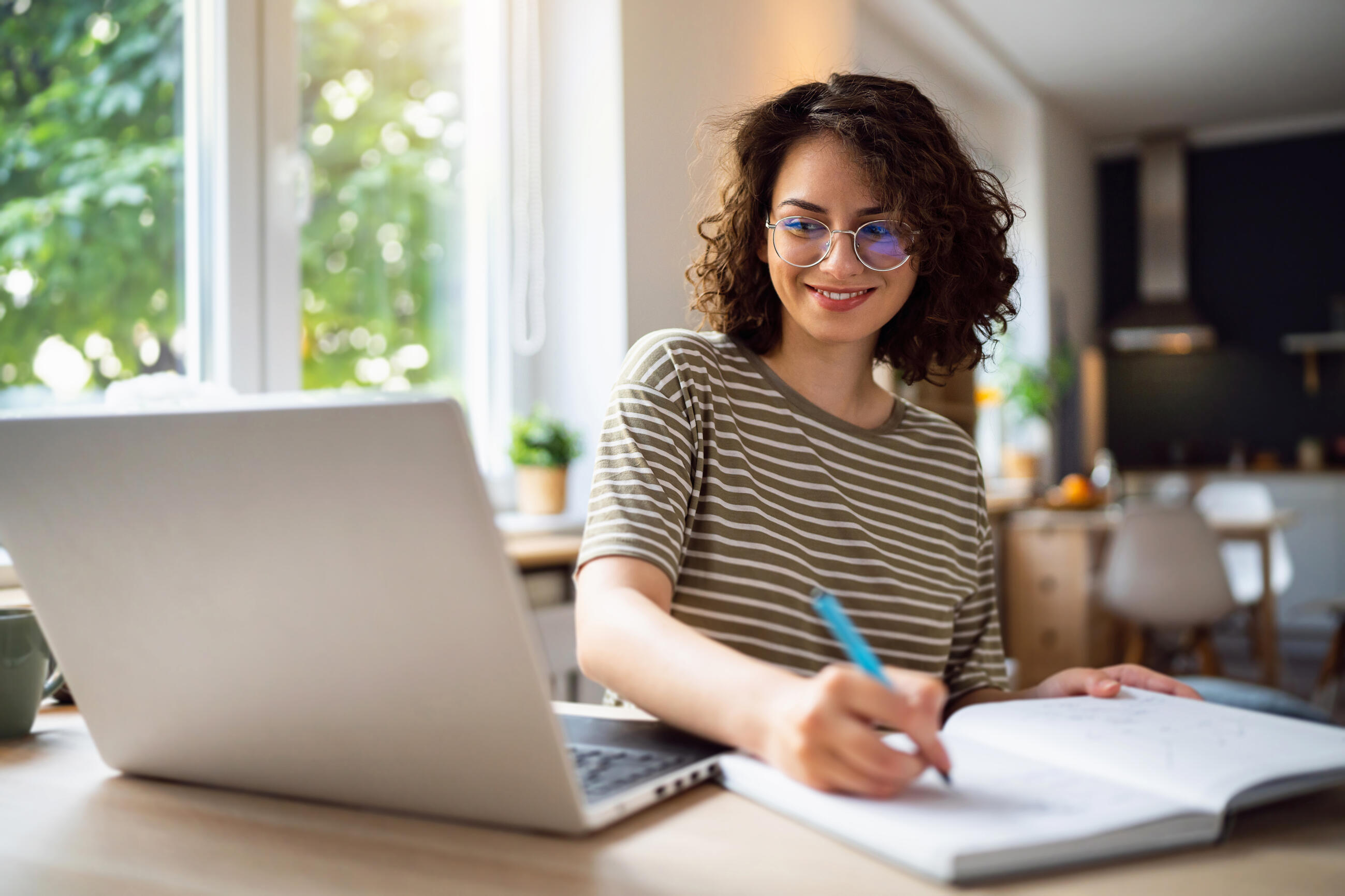 Woman sitting at laptop, writing something in a notebook with a pen